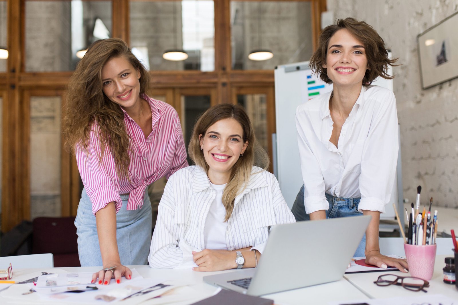 Group of young cheerful women with laptop happily looking in camera spending time together at work. Beautiful stylish girls joyfully working in modern cozy office