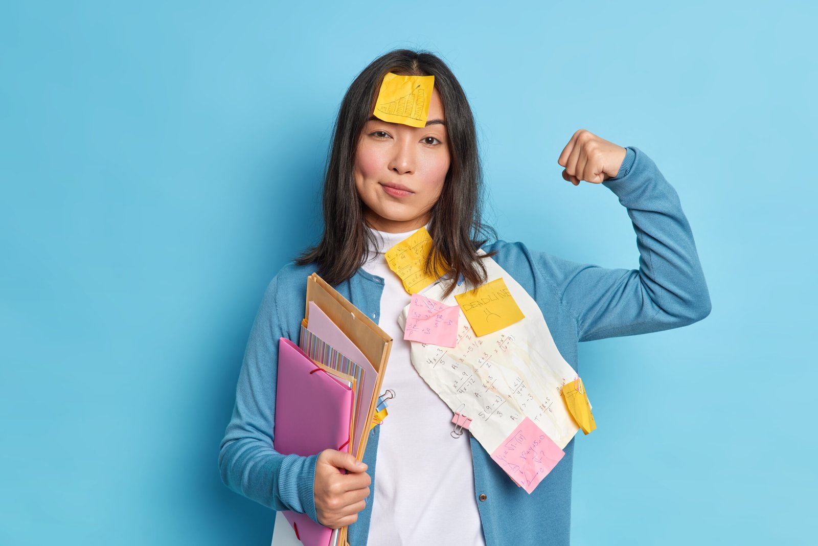 Powerful female student raises arm and shows muscles feels confident after working on diploma paper wears stickers on forehead holds folders isolated over isolated over blue studio background