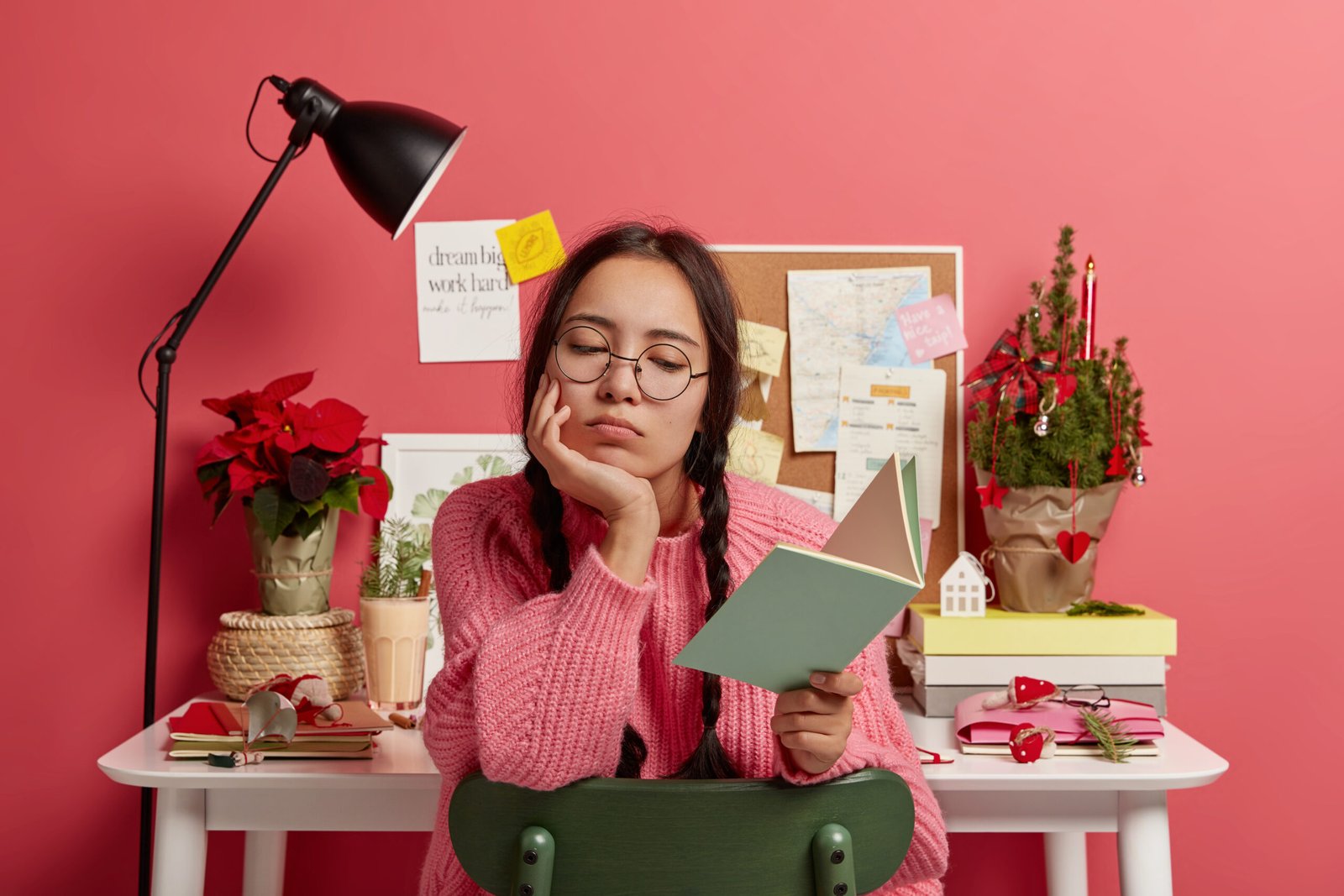 Serious Asian student learns information from textbook, does school homework, wears round glasses and sweater, poses against desktop, New Year tree, winter cocktail, board with sticky notes behind